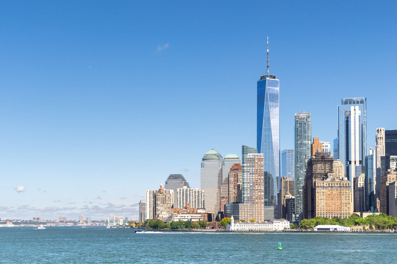 The skyline of New York City as seen from a distance with water in the foreground, and clear blue sky in the background.